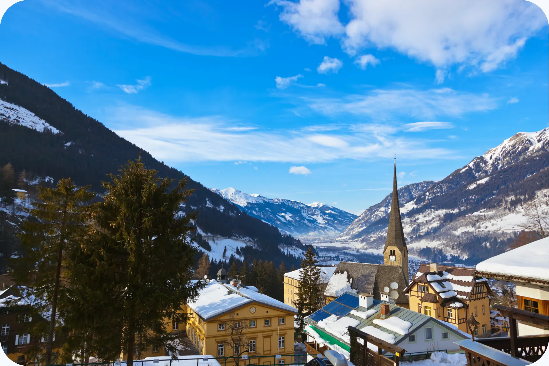 Winteruitzicht over Bad Gastein met besneeuwde daken, een kerktoren en bergen onder een heldere blauwe lucht.