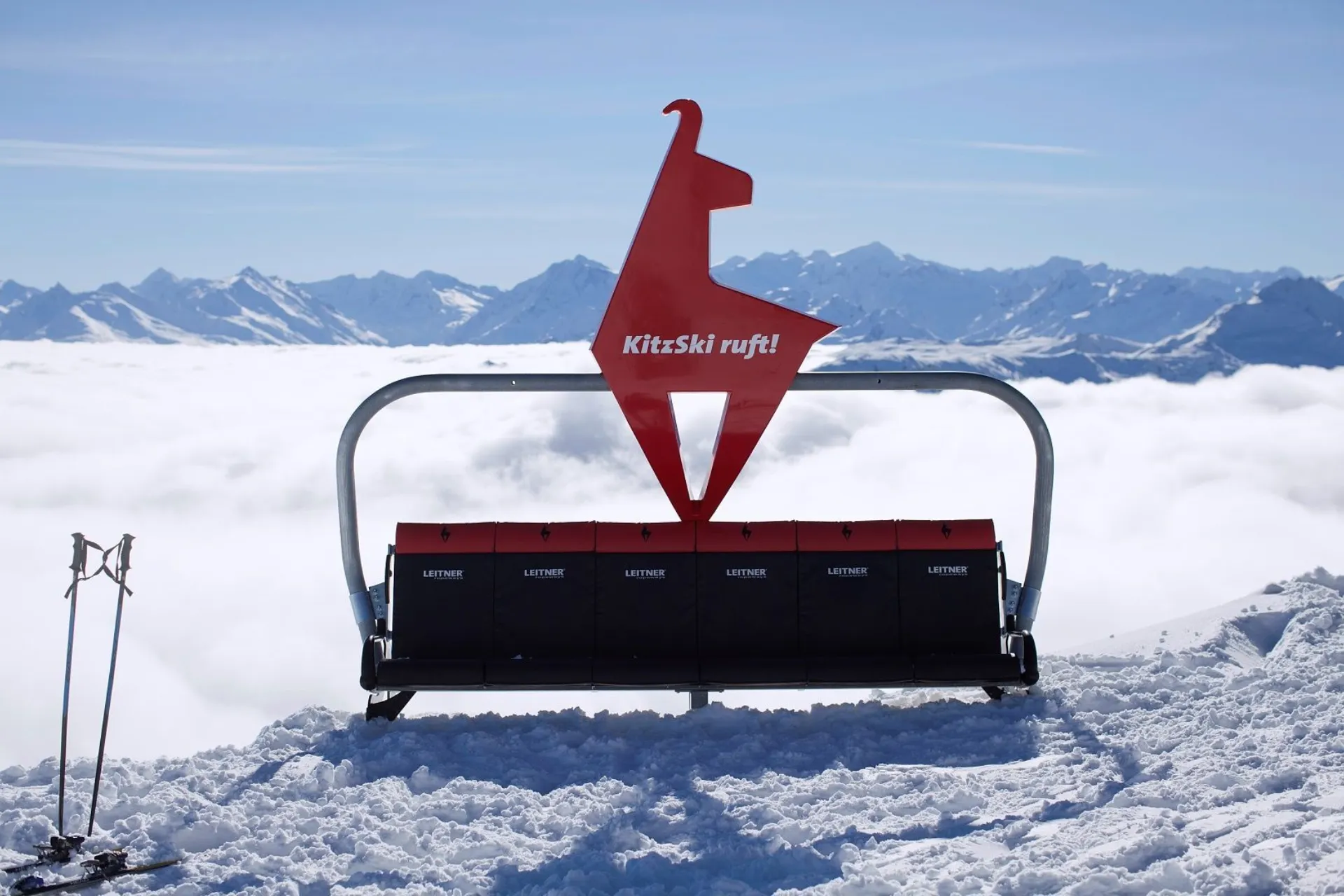 Bankje van een skilift in de sneeuw naast skistokken met een ''KitzSki ruft!'' bord.