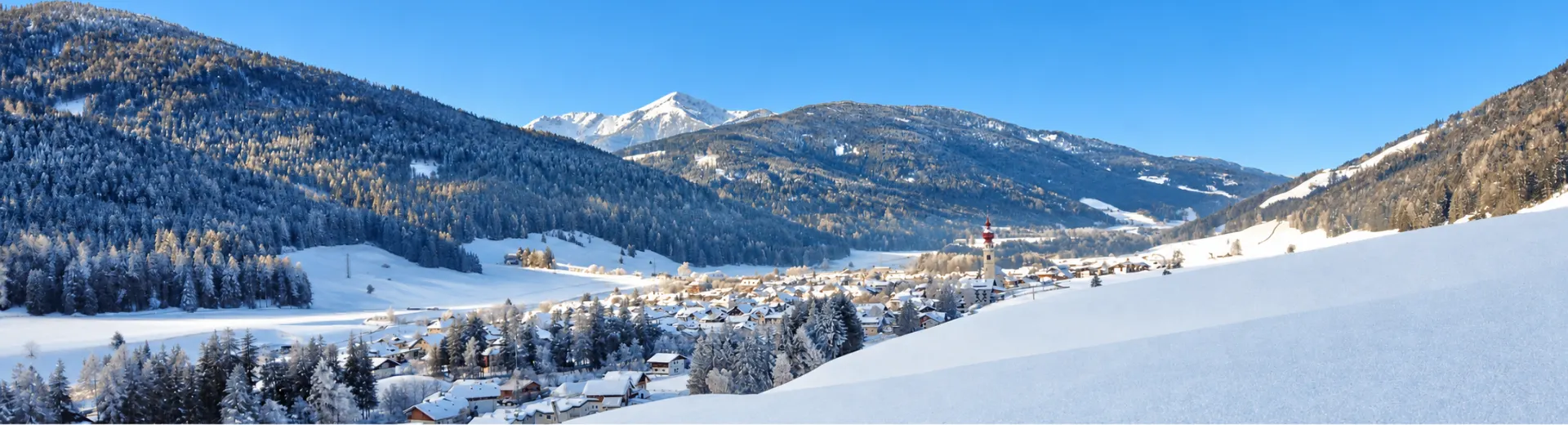 Zonnig winterlandschap met besneeuwd bergdorp, kerk en omliggende Alpen onder een strakblauwe lucht.