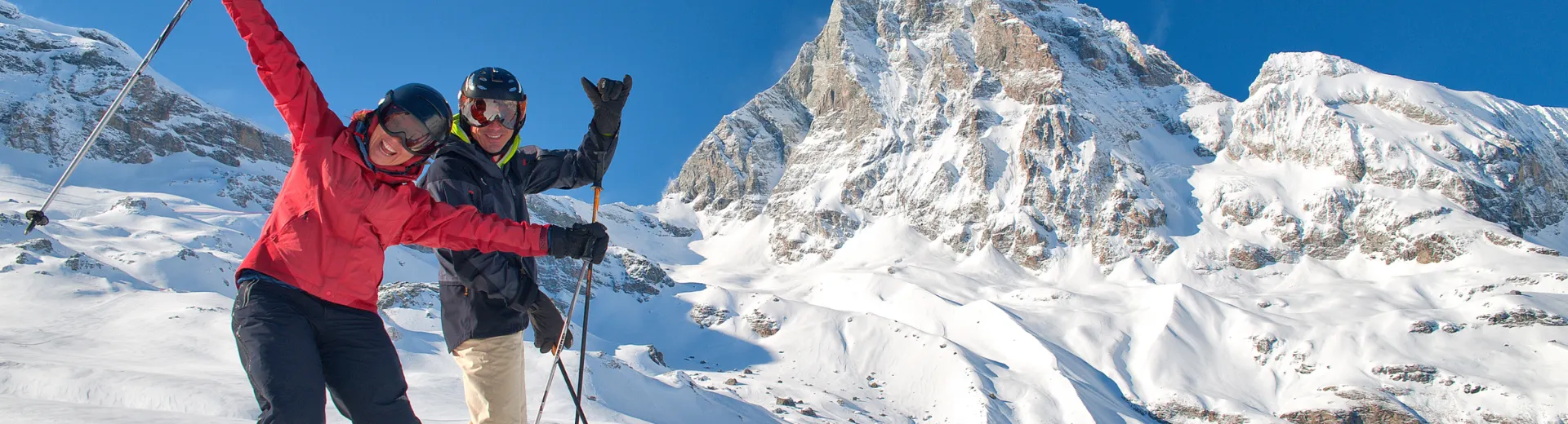 Twee skiërs juichen op een besneeuwde berg met indrukwekkende pieken op de achtergrond