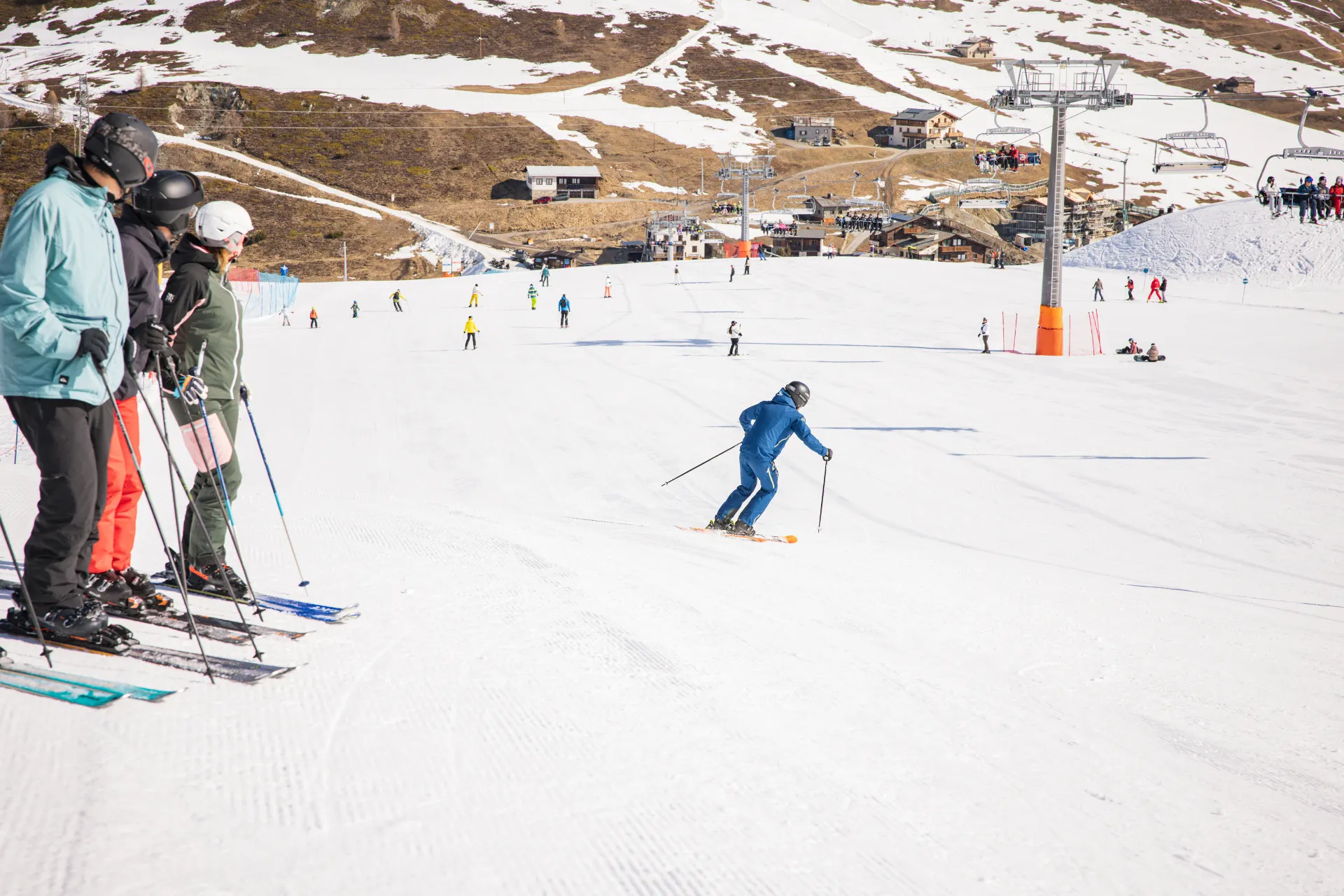 Skiërs op een brede piste in met skiliften, bergrestaurants en besneeuwde hellingen op de achtergrond.