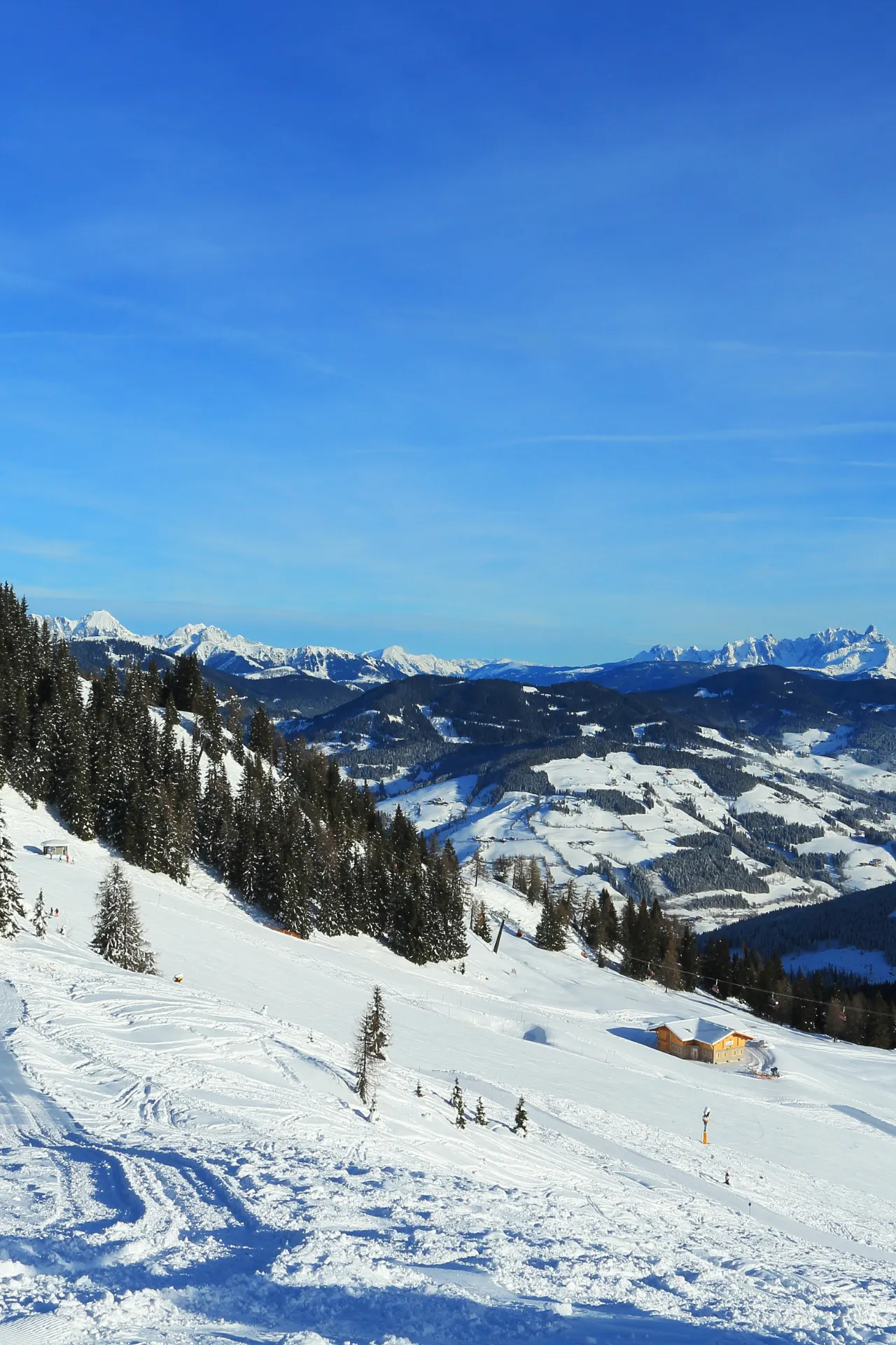 Een berg waar skisporen instaan met uitzicht over de bergen. Een houten huisje midden op de berg.